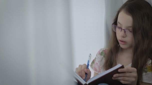 Красивая маленькая девочка в очках диктует себя и пишет в дневнике — стоковое видео