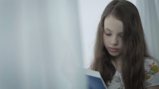Belle petite fille sérieuse lisant un livre par fenêtre . — Video