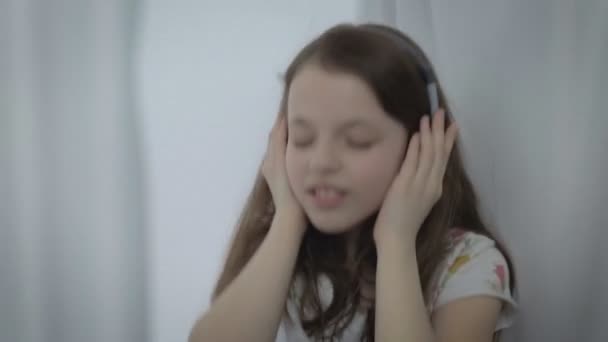 Mooie meisje in koptelefoon emotioneel een liedje zingen en dansen bij raam. — Stockvideo
