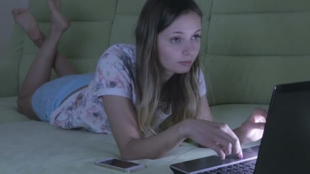 Mooi meisje liggend op Bank werken op zijn laptop. Ze beantwoordt de oproep op een smartphone. — Stockvideo