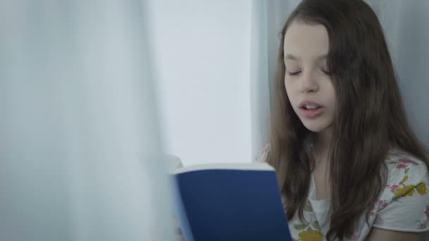 漂亮的小女孩读有趣的书，由窗口. — 图库视频影像