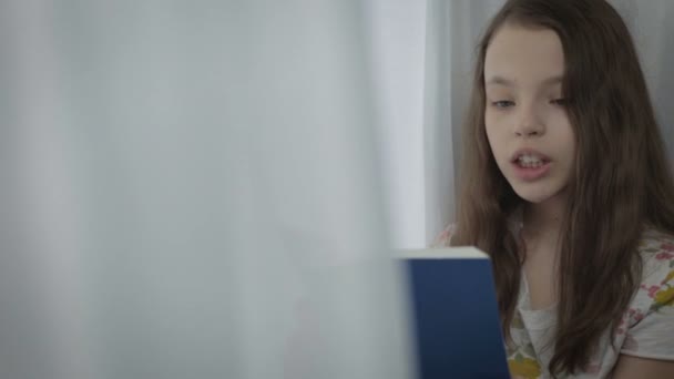 Όμορφο κοριτσάκι διαβάζοντας ένα ενδιαφέρον βιβλίο από το παράθυρο. — Αρχείο Βίντεο