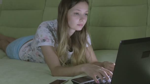 美丽的女孩躺在沙发上在他的笔记本电脑工作。她回答在智能手机上的呼叫. — 图库视频影像
