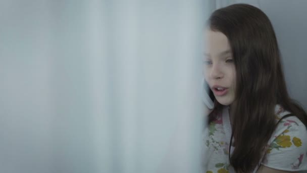 Красивая маленькая девочка эмоционально разговаривает по смартфону у окна . — стоковое видео