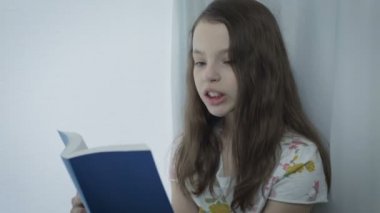 Güzel küçük kız pencere tarafından ilginç bir kitap okuma.