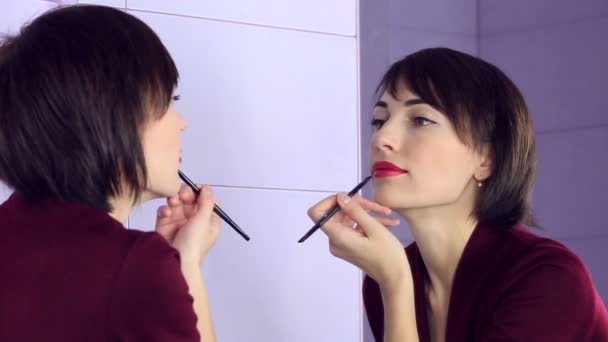 Mujer de belleza aplicando maquillaje. Hermosa chica mirando en el espejo y se pinta los labios con pincel . — Vídeos de Stock