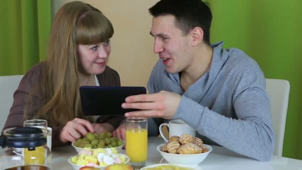 Junges fröhliches verliebtes Paar blickt auf Tablet und spricht beim Frühstück. — Stockvideo