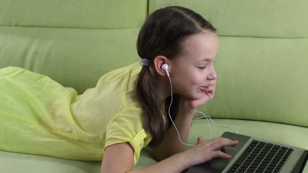 Красивая маленькая девочка лежит на диване и смотрит кино на ноутбуке . — стоковое видео