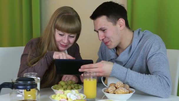 Jonge vrolijke paar verliefd op zoek op Tablet PC en praten over ontbijt. — Stockvideo