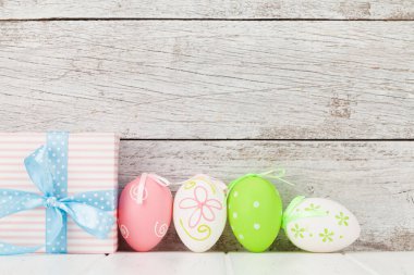 Ahşap duvarın önünde renkli paskalya yumurtaları ve hediye kutusuyla Paskalya tebrik kartı. Selamlarınız için yer var.