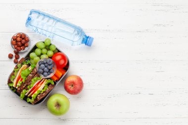 Sağlıklı beslenme çantası, sandviç ve taze sebzeler, su şişesi, fındık ve ahşap masada meyveler. Üst görünüm düzlüğü kopyalama alanı ile yatıyordu