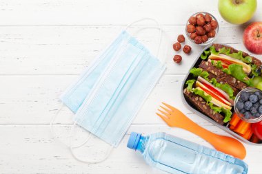 Tıbbi maske ve sağlıklı beslenme çantasıyla birlikte sandviç ve taze sebzeler, su şişesi, fındık ve meyveler masada. Üst görünüm düzlüğü kopyalama alanı ile yatıyordu