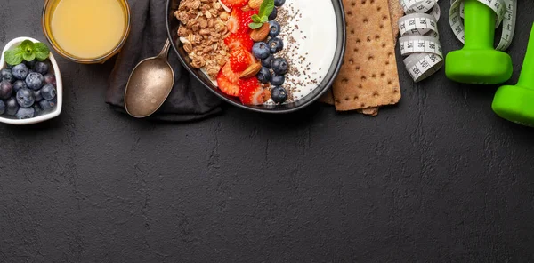 Healthy breakfast with bowl of granola, yogurt and fresh berries. Fitness protein meal. Dumbbells and tape measure. Diet concept. Top view flat lay with copy space