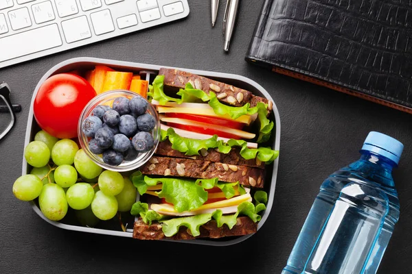 Healthy office lunch box with sandwich and fresh vegetables, water bottle, nuts and fruits on desk. Top view flat lay