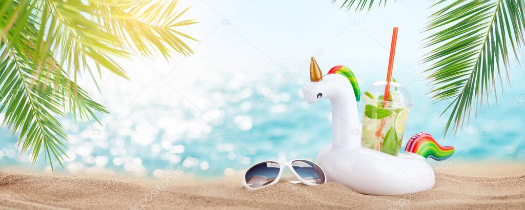 Fresh cold mojito cocktail in inflatable unicorn toy and sunglasses on tropical beach with palms and bright sand. Summer sea vacation and travel concept