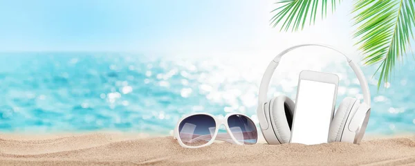Smartphone, sunglasses and headphones on tropical sea beach with palms and bright sand. Summer sea vacation, music and travel concept. With blank screen for your app