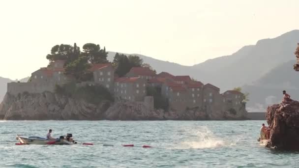 Gente saltando al mar con la famosa isla de Sveti Stefan al fondo — Vídeos de Stock