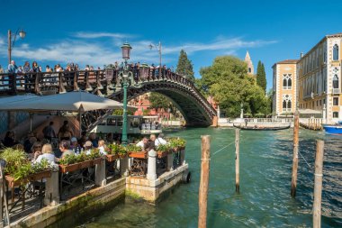 Venedik, İtalya - 11 Ekim 2024: Turistler Büyük Kanal üzerindeki ahşap Accademia Köprüsü 'nde yürürken, diğerleri güneşli bir günde deniz kenarındaki bir restoran terasında yemeğin tadını çıkarıyorlar..