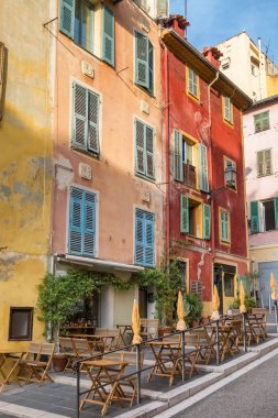 Güzel, France. Tarihi merkezde mavi panjurlu canlı sarı ve kırmızı Akdeniz cephelerine karşı ahşap sandalyeli ve masalı rahat bir açık hava restoranı terası..