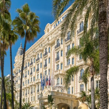 Cannes, France - September 21, 2024: Low angle view of the majestic Carlton Hotel facade and flags framed by tall palm trees under a blue sky on the Cote d Azur.