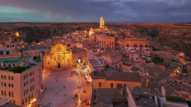 Matera 'nın mavi saatte çekilmiş robot görüntüleri tarih öncesi Sassi mağara evlerini ve Roma katedralini gösteriyor. Aydınlatılmış UNESCO şehri Basilicata İtalya 'da alacakaranlık gökyüzünün altında parlıyor.