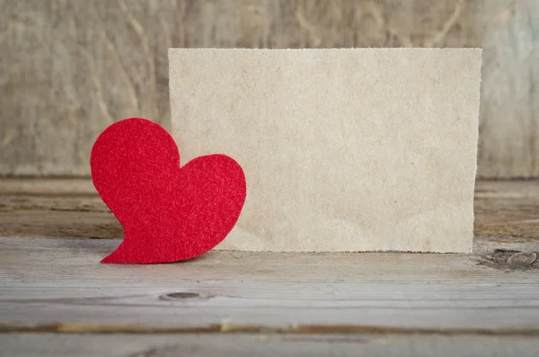 Red fabric heart with sheet of paper standing on a wooden board. — Stock Photo, Image