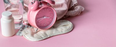Alarm saati, göz maskesi, haplar ve pembe arka planda bir bardak su.