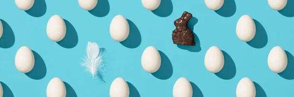 Patrón hecho de huevos blancos, banny chocolate y pluma de pájaro sobre fondo azul, bandera —  Fotos de Stock