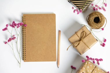 Kraft kağıdına sarılı hediye kutusu, beyaz arka planda not defteri ve pembe çiçekler. Düz yatış stili.