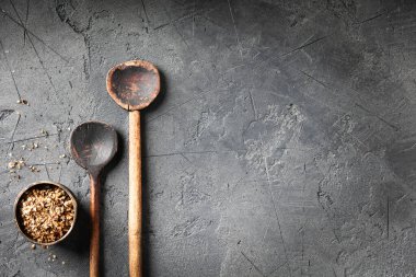 Koyu gri bir taş yüzeyde, küçük bir kase ezilmiş baharatın yanında iki kırsal tahta kaşık. Mutfak, köy mutfağı veya yemek blogu temaları için ideal.