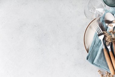 Mavi keten peçeteli seramik bir tabağın, ahşap saplı çatal bıçak takımının ve hafif desenli bir arka planda cam süslemenin bulunduğu minimal ve zarif bir masa ortamının üst görüntüsü. Kurumuş çiçekler kırsal bir dokunuş ekler. Yemek, yaşam tarzı ya da misafirperverlik için mükemmel.