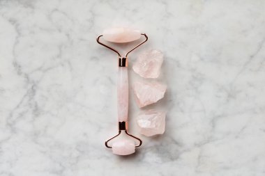 Pembe bir gül kuvars yüz silindiri, saf gül kuvars taşlarının yanında mermer bir zemin üzerinde, güzelliği ve iyileşmeyi simgeliyor..