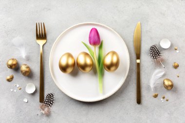 Beyaz tabakta altın yumurtalar, pembe lale, altın çatal bıçak ve hafif taşlı arka planda tüylerle süslü Paskalya kompozisyonu. Kopyalama alanı olan minimal şenlik düzlüğü.