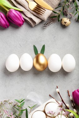 Beyaz ve altın yumurtalarla süslü Paskalya dairesi, pembe laleler, bahar yeşilliği ve hafif taşlı arka planda altın çatal bıçak seti. Kopya alanı ile modern festival konsepti.