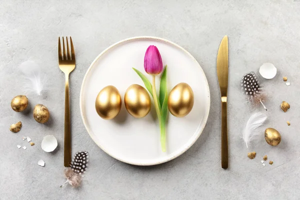 Beyaz tabakta altın yumurtalar, pembe lale, altın çatal bıçak ve hafif taşlı arka planda tüylerle süslü Paskalya kompozisyonu. Kopyalama alanı olan minimal şenlik düzlüğü.