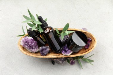 Zenginlik temalı bir daire; kehribar camı, gerekli yağ şişeleri, ham ve cilalanmış ametist kristalleri, okaliptüs dalları ve hafif bir taş zemin üzerinde, rahatlatıcı, bütünsel bir titreşim için ahşap bir kasede düzenlenmiş bir damlacıkla uzanır..