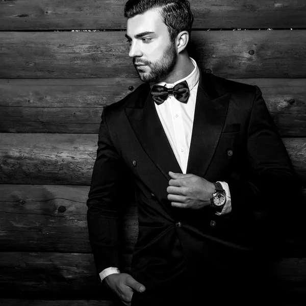 Ritratto di giovane bell'uomo alla moda contro parete di legno In completo nero e papillon. Foto di moda in bianco e nero sullo sfondo in legno . — Foto Stock