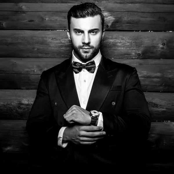 Portret van jonge mooie modieuze man tegen houten muur In zwart pak & strikje. Zwart-wit mode foto tegen houten achtergrond. — Stockfoto