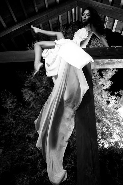Attractive girl in white long dress poses on wooden beam. Black-white outdoor photo.