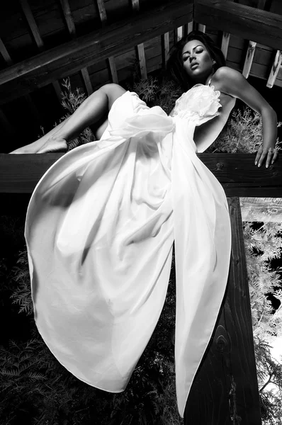 Jolie fille en blanc robe longue pose sur poutre en bois. Photo extérieure noir-blanc . — Photo