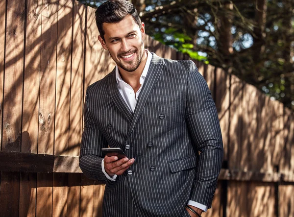 Homem bonito elegante em terno cinza clássico posa perto de cerca de madeira — Fotografia de Stock