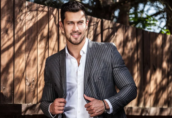 Elegant handsome man in classical grey suit poses near wooden fence — Stock Photo, Image