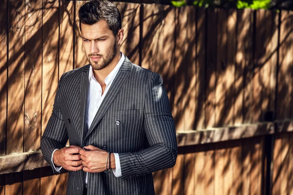 Elegante bell'uomo in classico abito grigio posa vicino alla recinzione in legno — Foto Stock