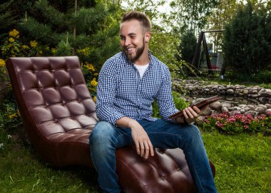 Young handsome man in casual clothes sit in luxury sofa with iPad in summer garden.