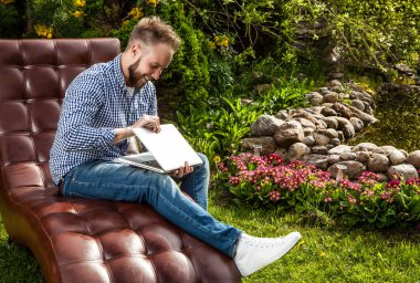Young handsome man in casual clothes sit in luxury sofa with notebook in summer garden.