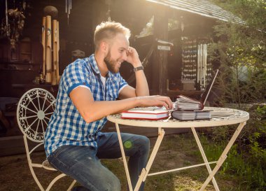 Young smiling handsome man in casual clothes work at an iron table with computer against country garden.
