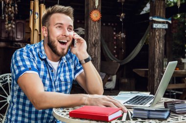 Young smiling handsome man in casual clothes work at an iron table with computer against country garden.