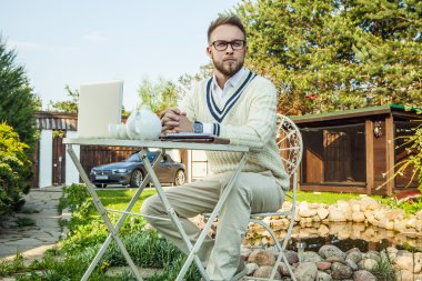 Young stylish handsome man in bright confidential clothes work at iron table with computer & teapot against country garden.