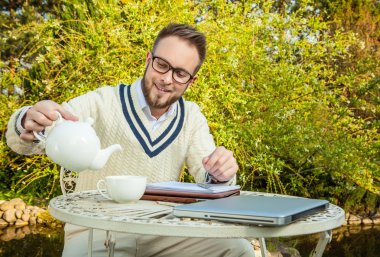 Young stylish handsome man in bright confidential clothes work at iron table with computer & teapot against country garden.