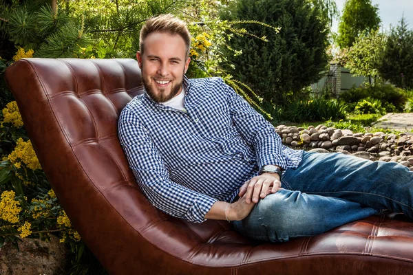 Joven hombre guapo en ropa casual sentarse en un sofá de lujo en el jardín de verano . — Foto de Stock